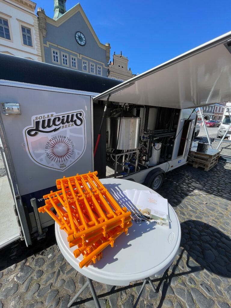 Mobile beer truck with side open to reveal stainless brewing equipment, orange plastic racks on a round table in foreground, and a historic square in the background.